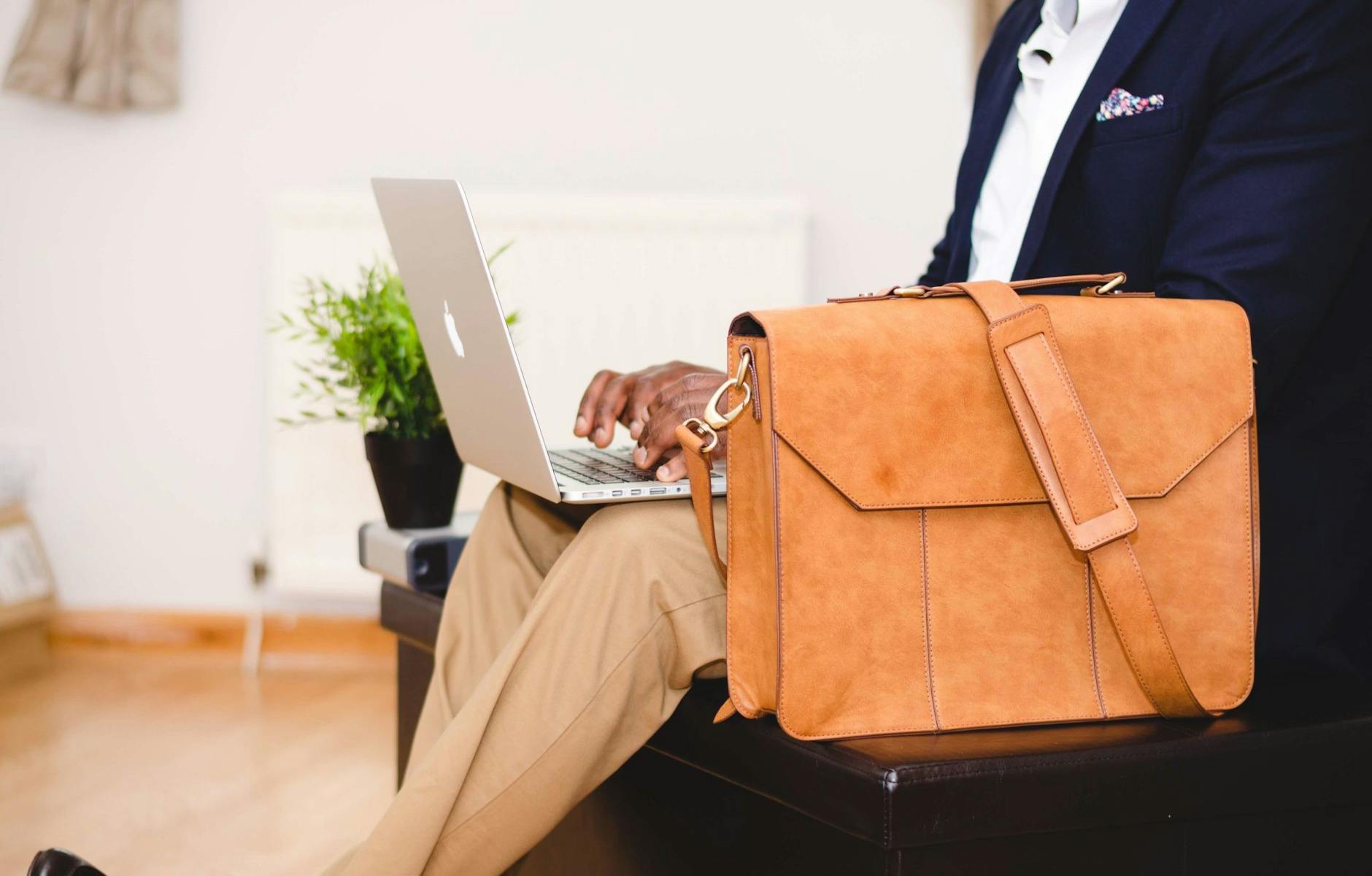 person wearing blue suit beside crossbody bag and using macbook